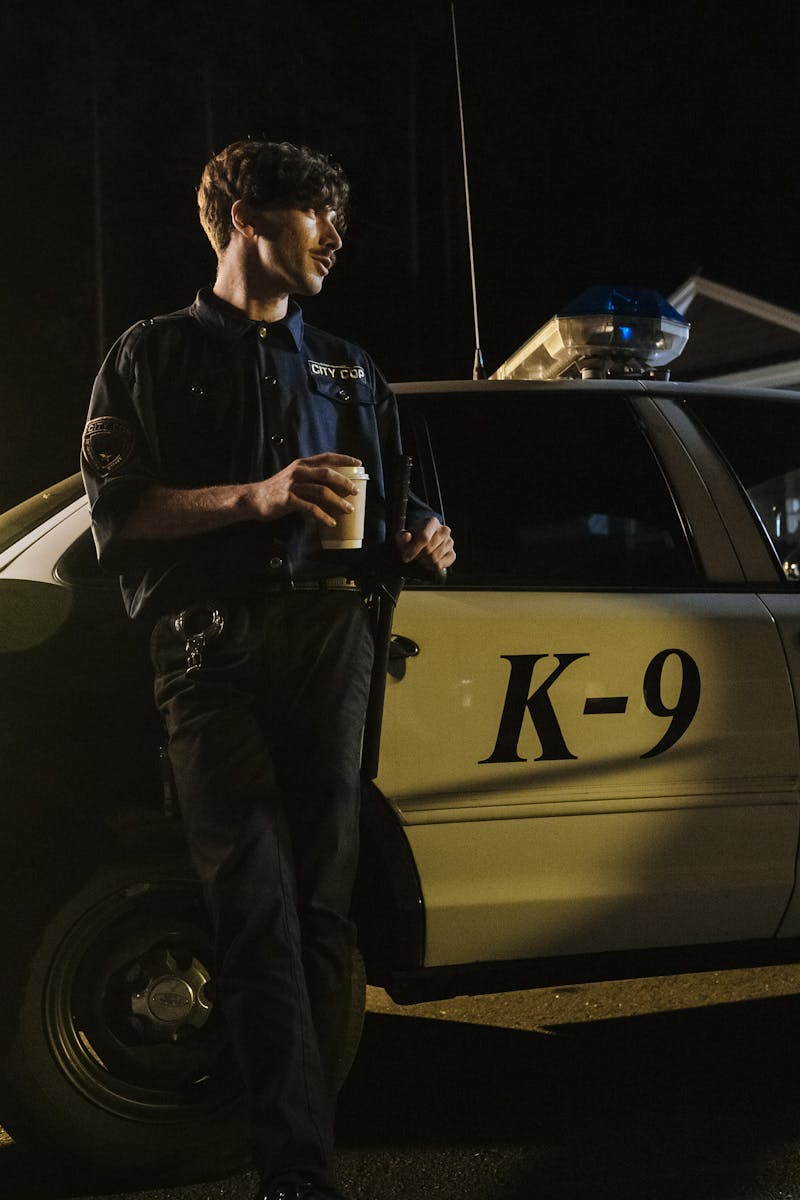 A police officer stands by a K-9 unit car at night, holding a coffee, illuminated by streetlights.