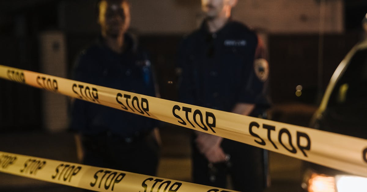 Two police officers standing behind a stop tape at night, scene partially illuminated by car headlights.