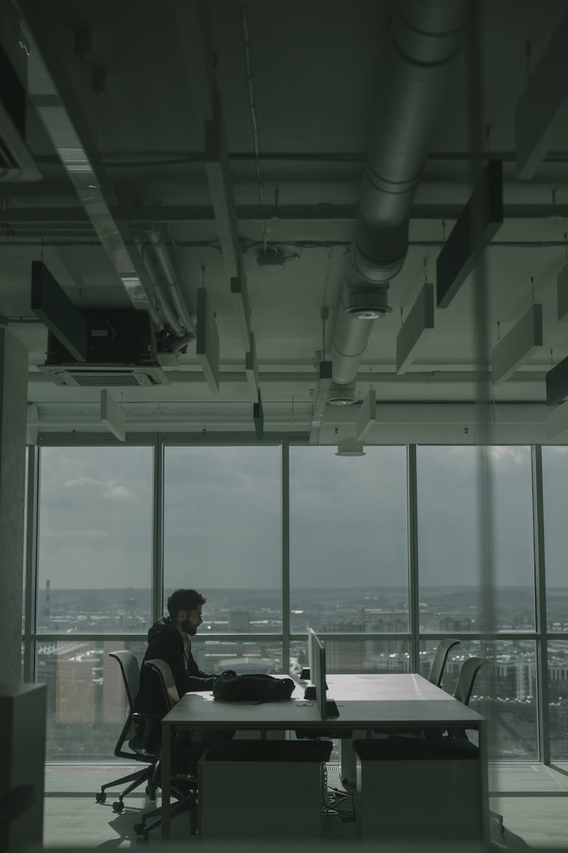 Man working on laptop in a modern office with expansive city views through large windows.