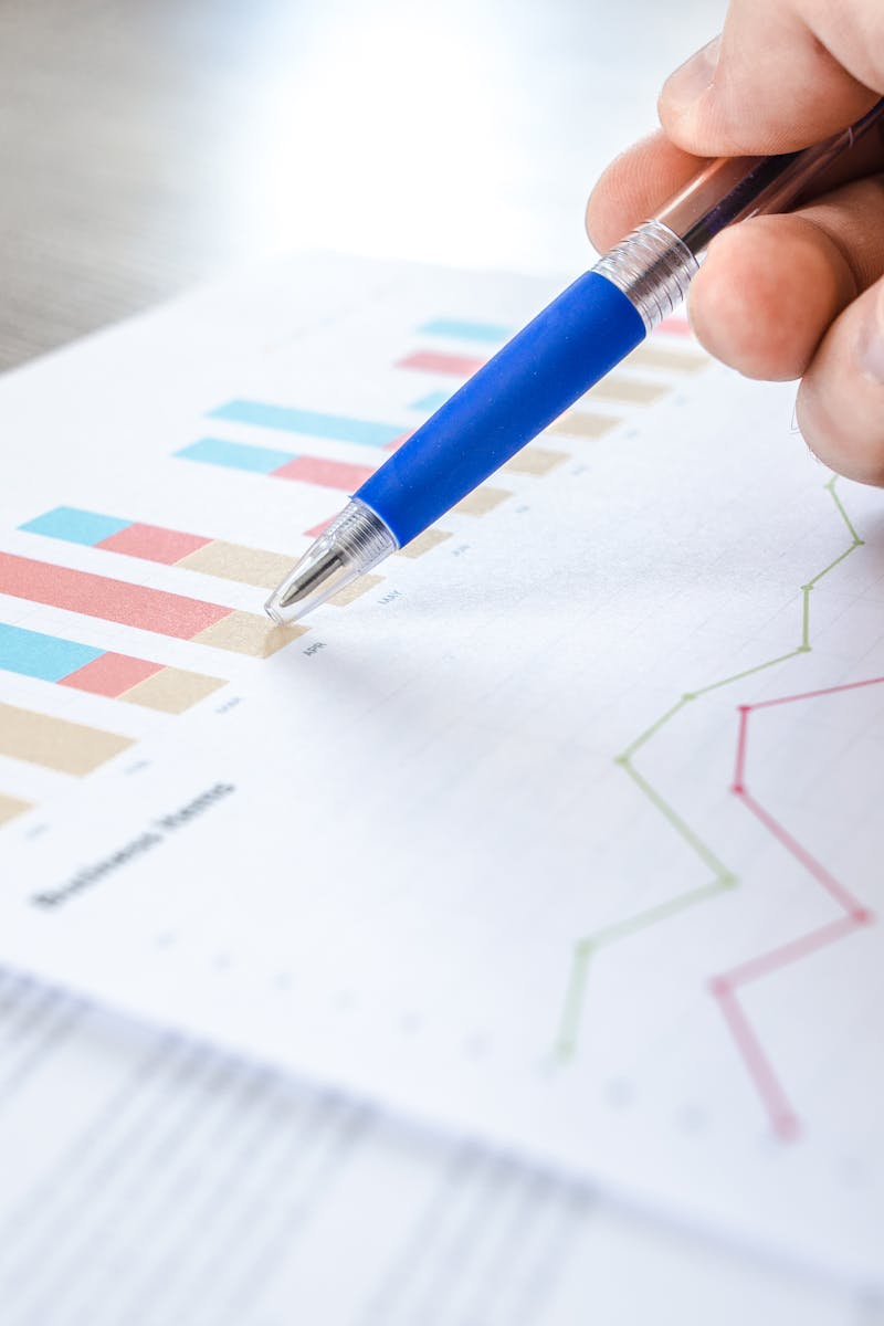 A close-up of a hand with a pen analyzing data on colorful bar and line charts on paper.