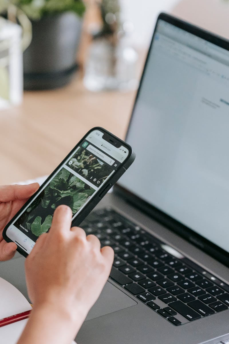 Person using a smartphone and laptop at a desk with coffee and plants, epitomizing modern remote work.